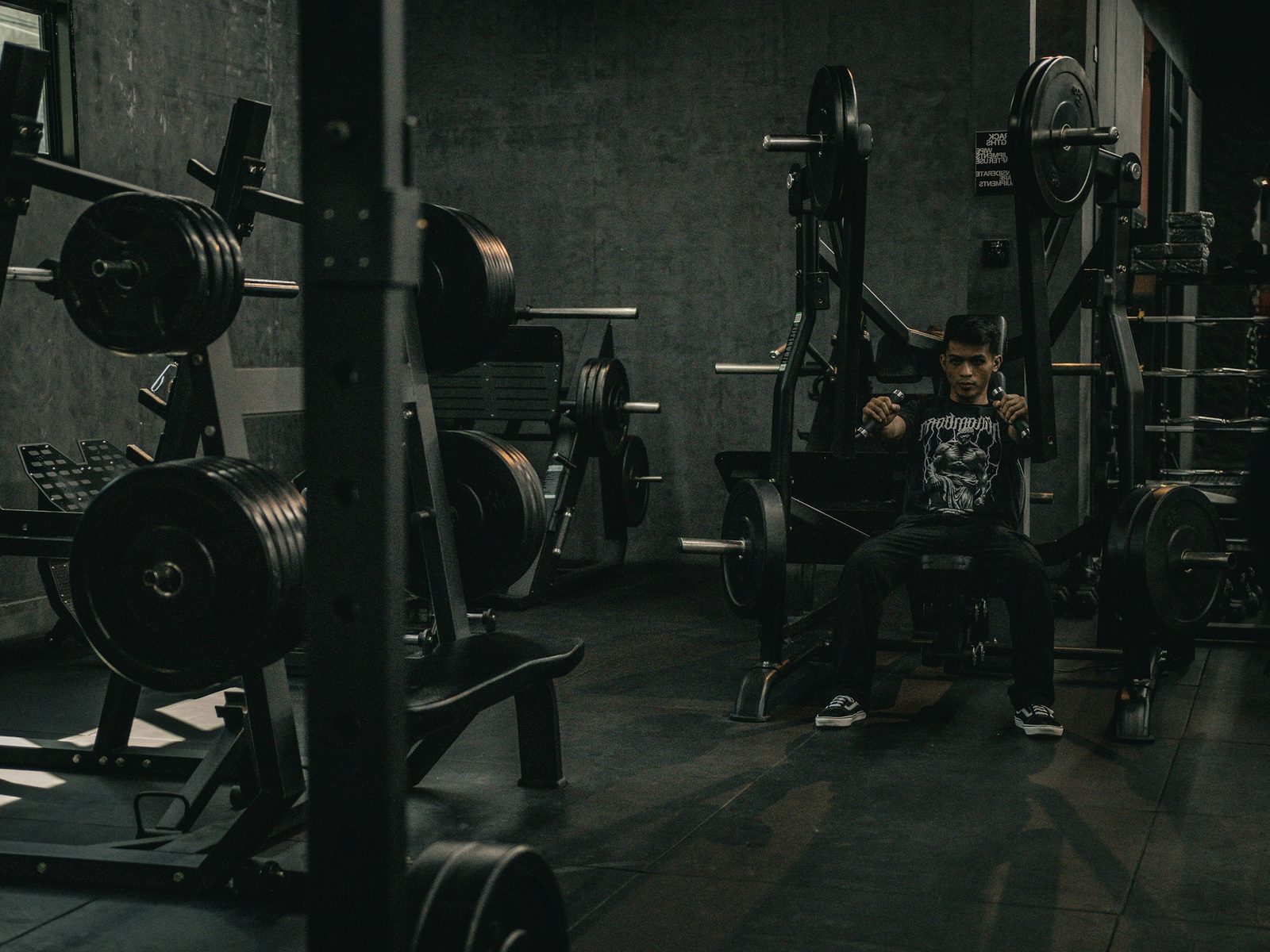 Sala de musculação vazia ao fim do dia, com um aluno treinando ao fundo